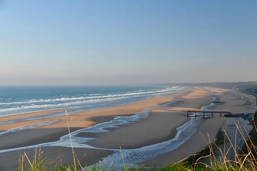 Vue panoramique d'une large plage de sable avec des vagues de l'océan, des bancs de sable et une passerelle, rappelant les plages du Débarquement en Normandie.