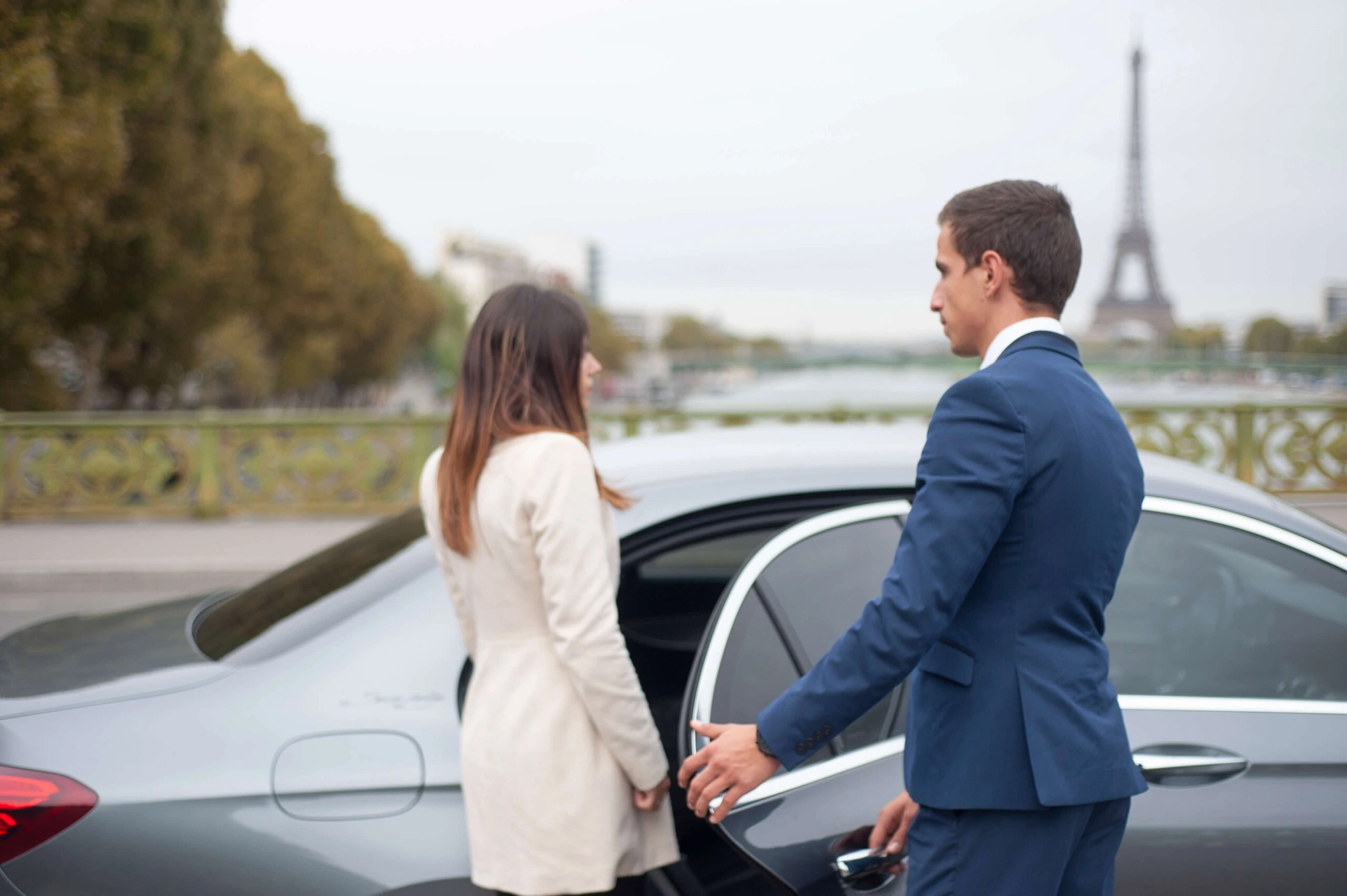 Chauffeur Service Prestige en costume ouvrant la porte d'une berline pour une cliente, avec la Tour Eiffel en arrière-plan à Paris.